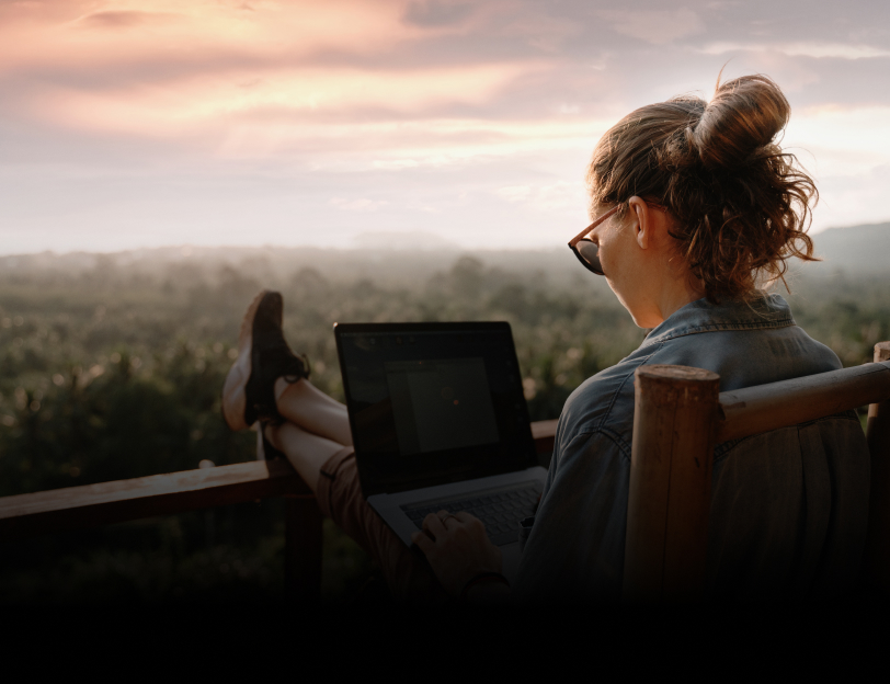  A woman sitting on a balcony overlooking a forest and working on a laptop.